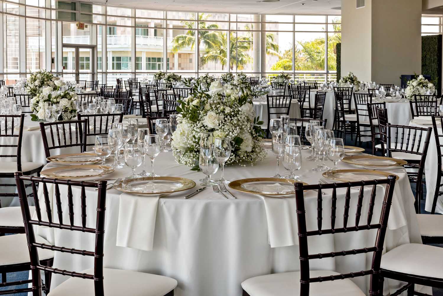 Formal reception setup in the Palm Ballroom with white florals, gold-rimmed chargers, and views of palm trees through floor-to-ceiling windows