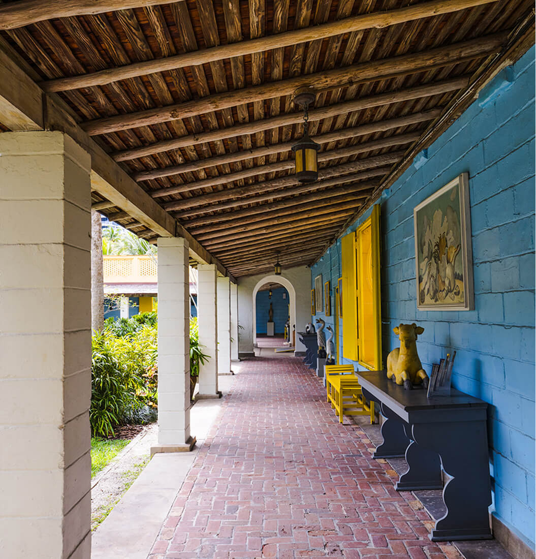 Covered loggia at Bonnet House with a bright blue wall, yellow shutters, herringbone brick floor, and garden beyond white columns
