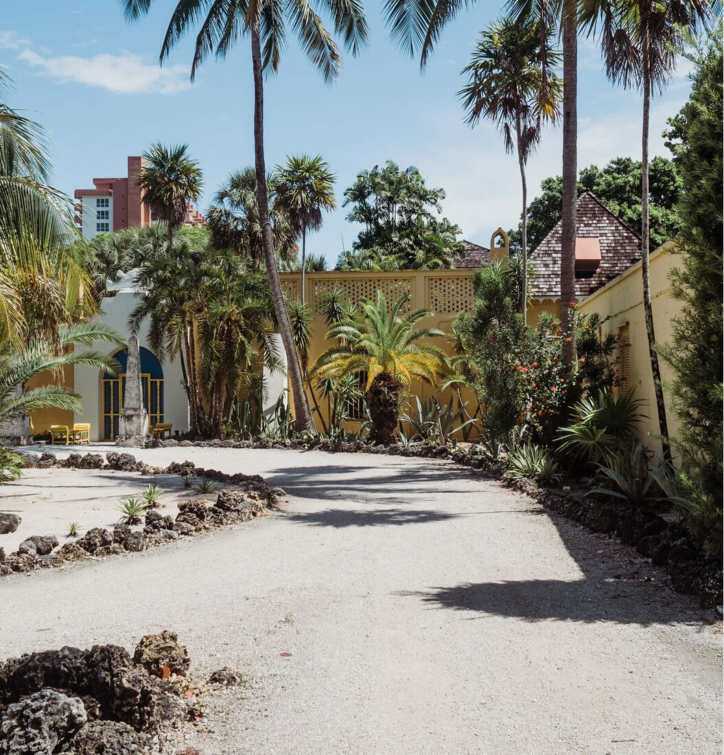 Sunny courtyard at Bonnet House with palm trees, a white arched doorway, yellow chairs, and a stone obelisk