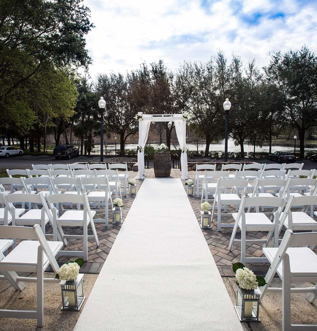 Outdoor lakeside wedding ceremony at Venue on Lake Lily in Maitland with white arch, aisle runner, folding chairs, and Lake Lily fountain in the background